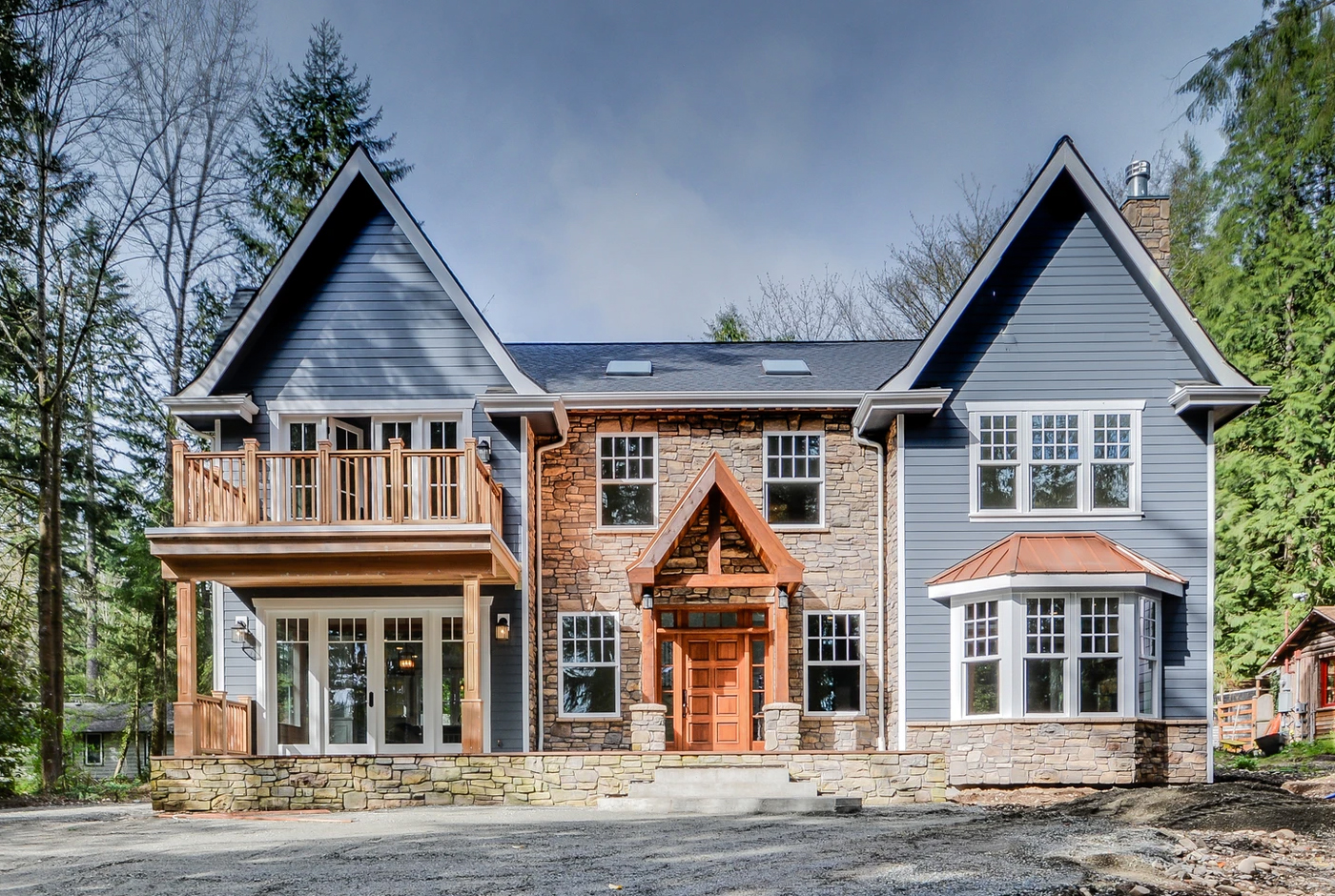 Two-story house with blue siding, stone accents, large windows, and a wooden front door. Features a balcony on the left and bay windows on both sides, surrounded by trees and a partially cloudy sky.