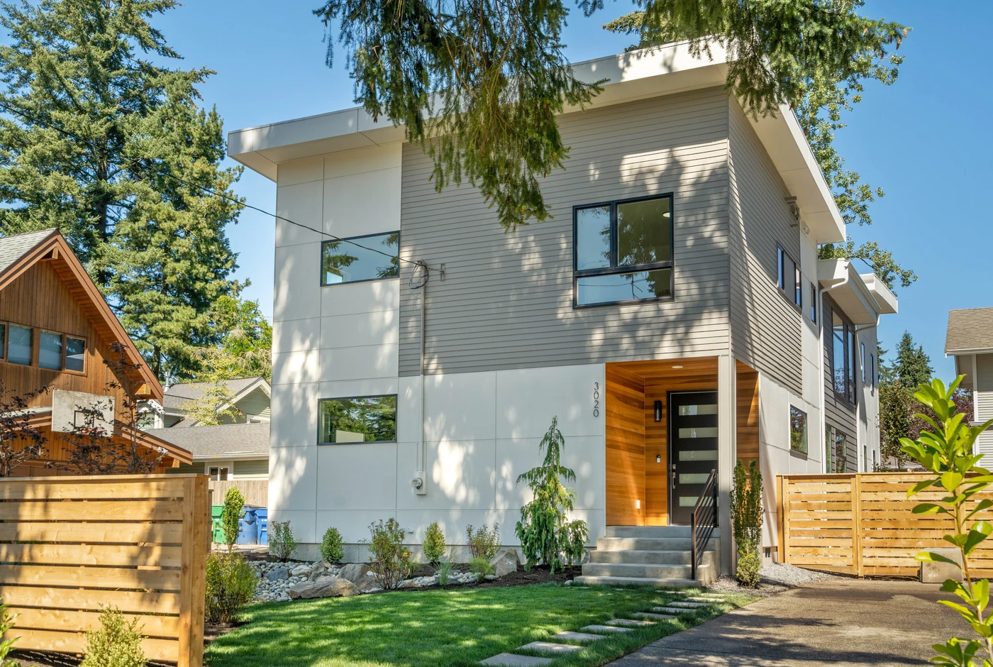 Modern two-story house with large windows, light gray siding, white panel accents, and a wooden entryway. The front yard features green grass, small shrubs, and a wooden fence.