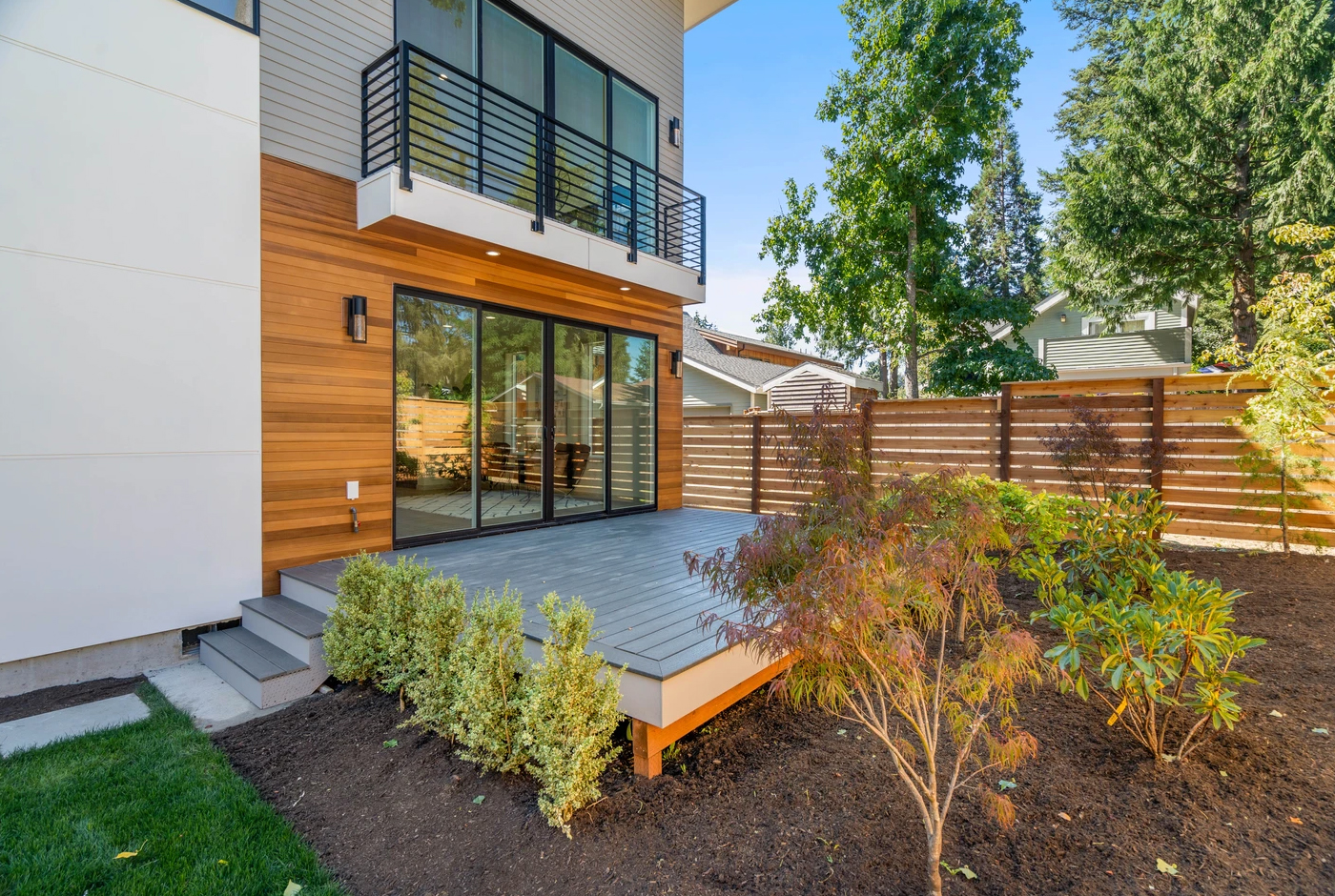 Modern house exterior with wooden siding, large sliding glass doors, a gray patio deck, shrubs along the edge, and a wooden fence surrounding a landscaped backyard with trees and mulch.