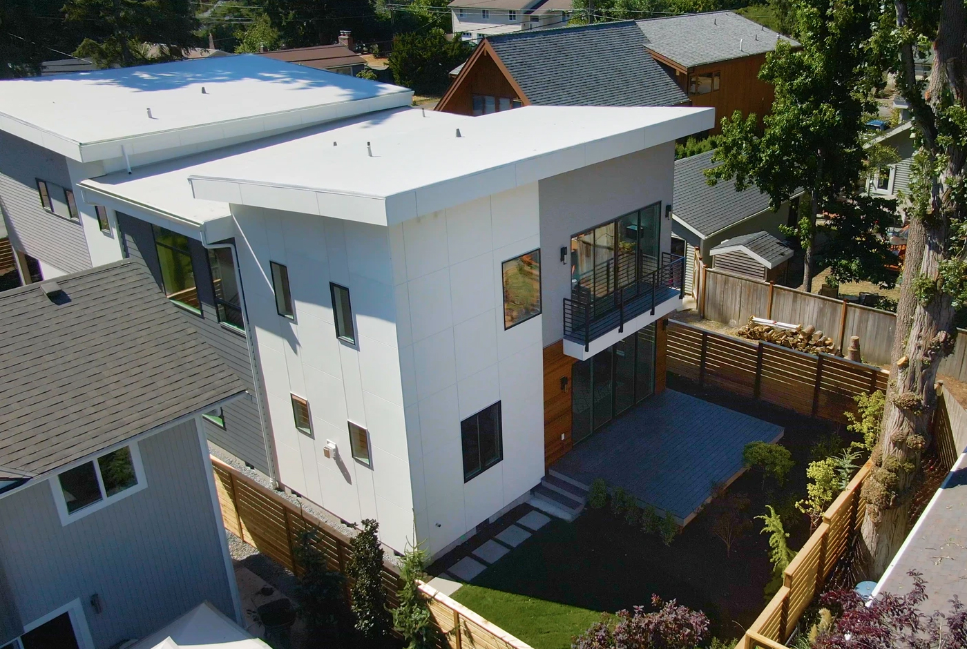 Modern two-story white house with a flat roof, large windows, and a balcony, surrounded by a wooden fence and landscaped yard, seen from an elevated perspective.