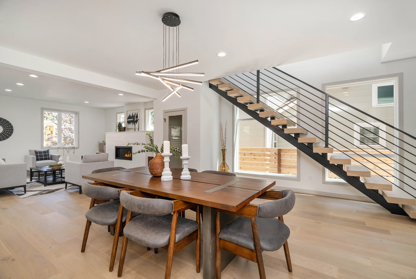 Modern dining area with a wooden table, gray chairs, unique chandelier, open staircase with metal railing, and view of a bright living room and large windows.