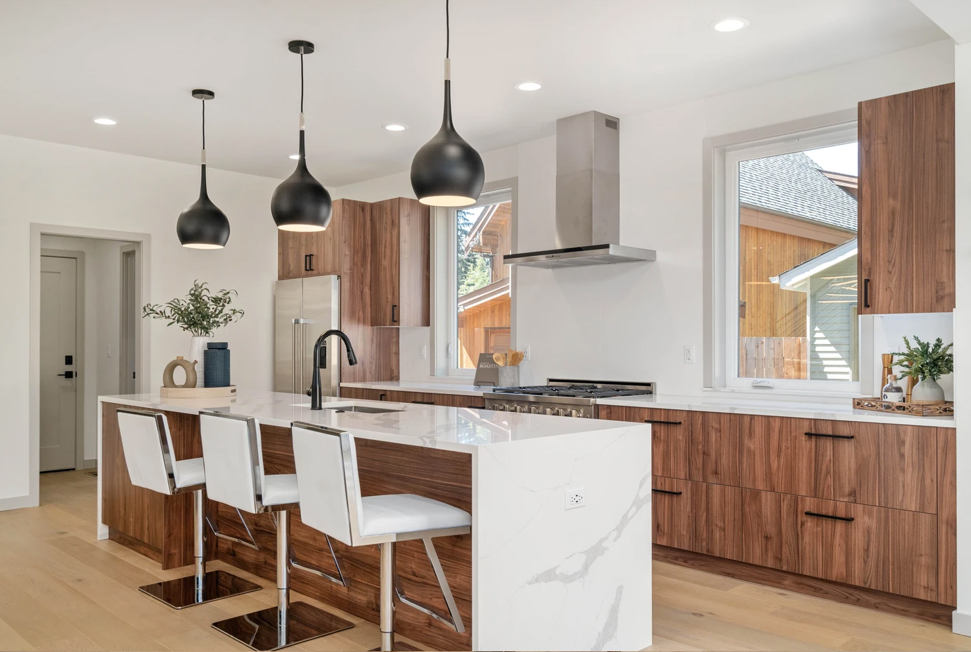 Modern kitchen with wood cabinets, white marble island, three white barstools, black pendant lights, stainless steel appliances, and large windows letting in natural light.