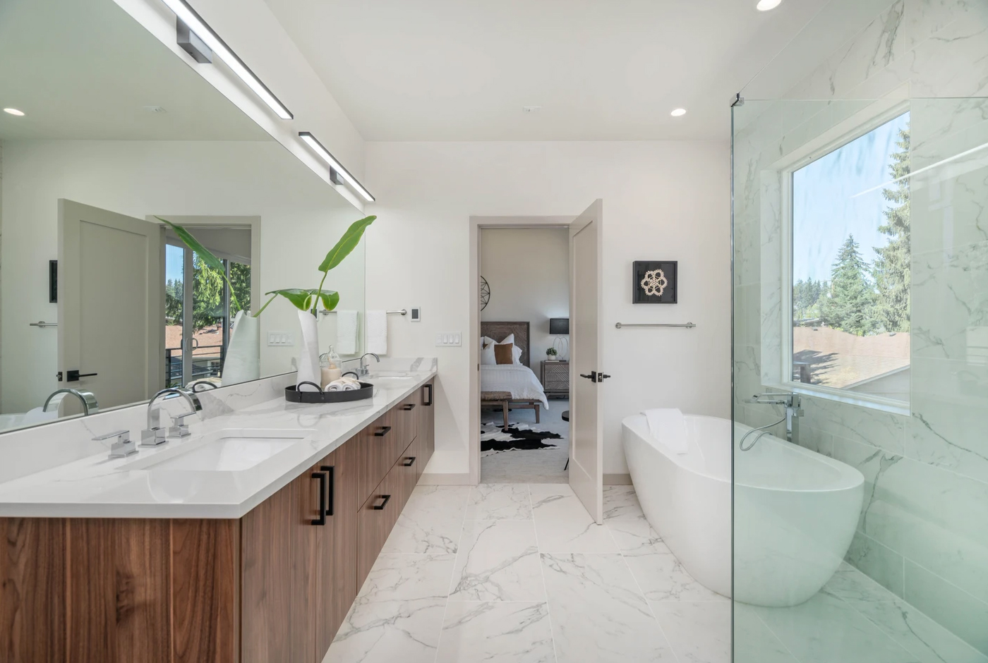 Modern bathroom with a double-sink vanity, large mirror, potted plant, glass shower, and freestanding bathtub. White marble floors and walls. Open door leads to a bedroom with a bed and cowhide rug. Large window with outdoor view.