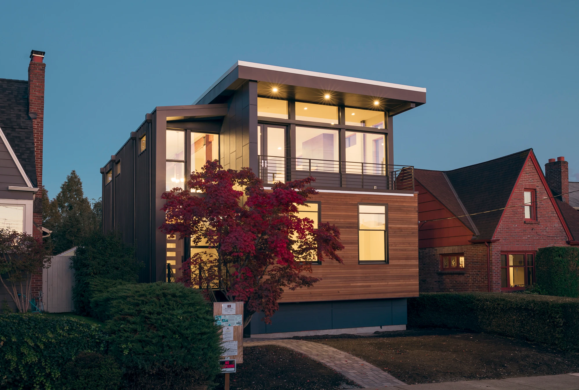 Modern two-story house with large windows and wood siding, illuminated at dusk. A small tree with red leaves stands in front, and neighboring houses are visible on either side.