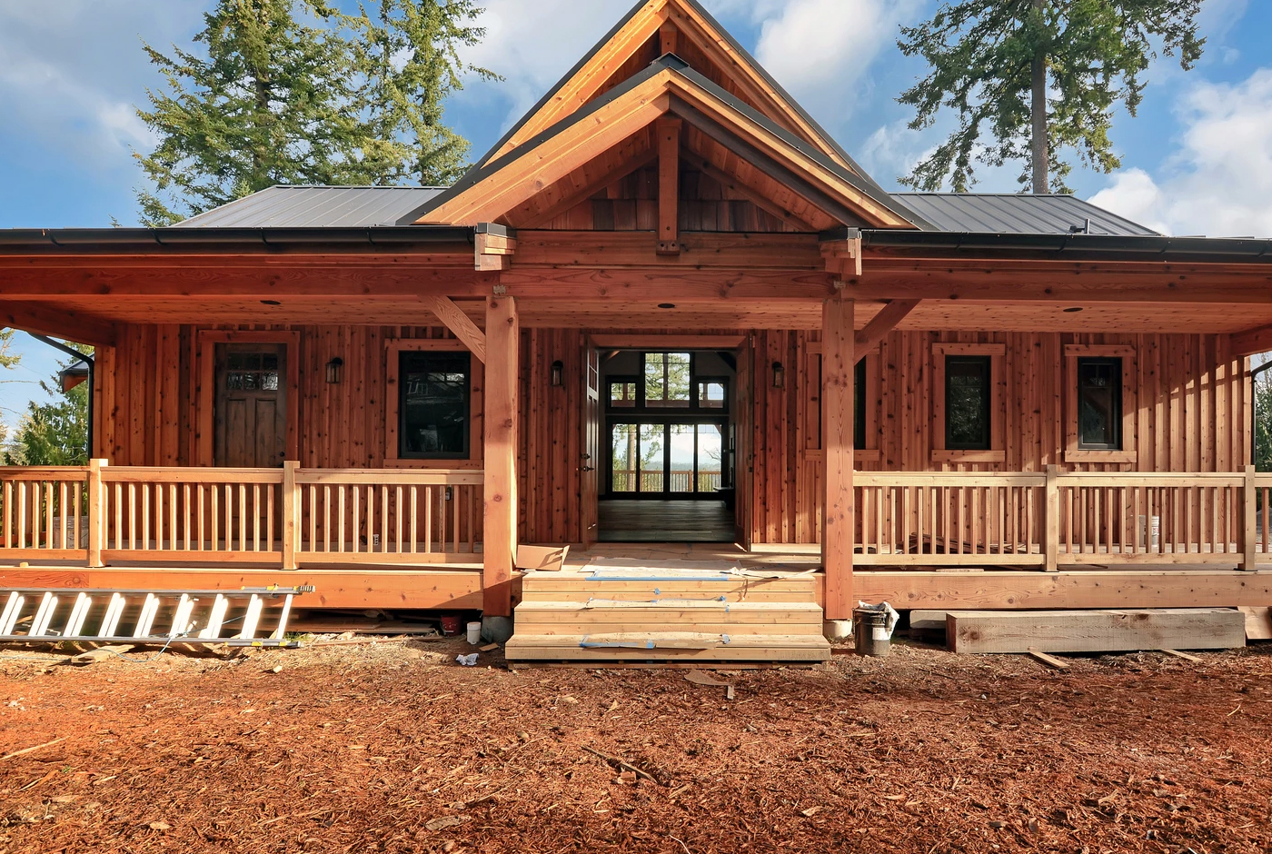 A wooden cabin with a covered front porch, steps leading up, several windows, and trees in the background. A ladder lies on the ground to the left.