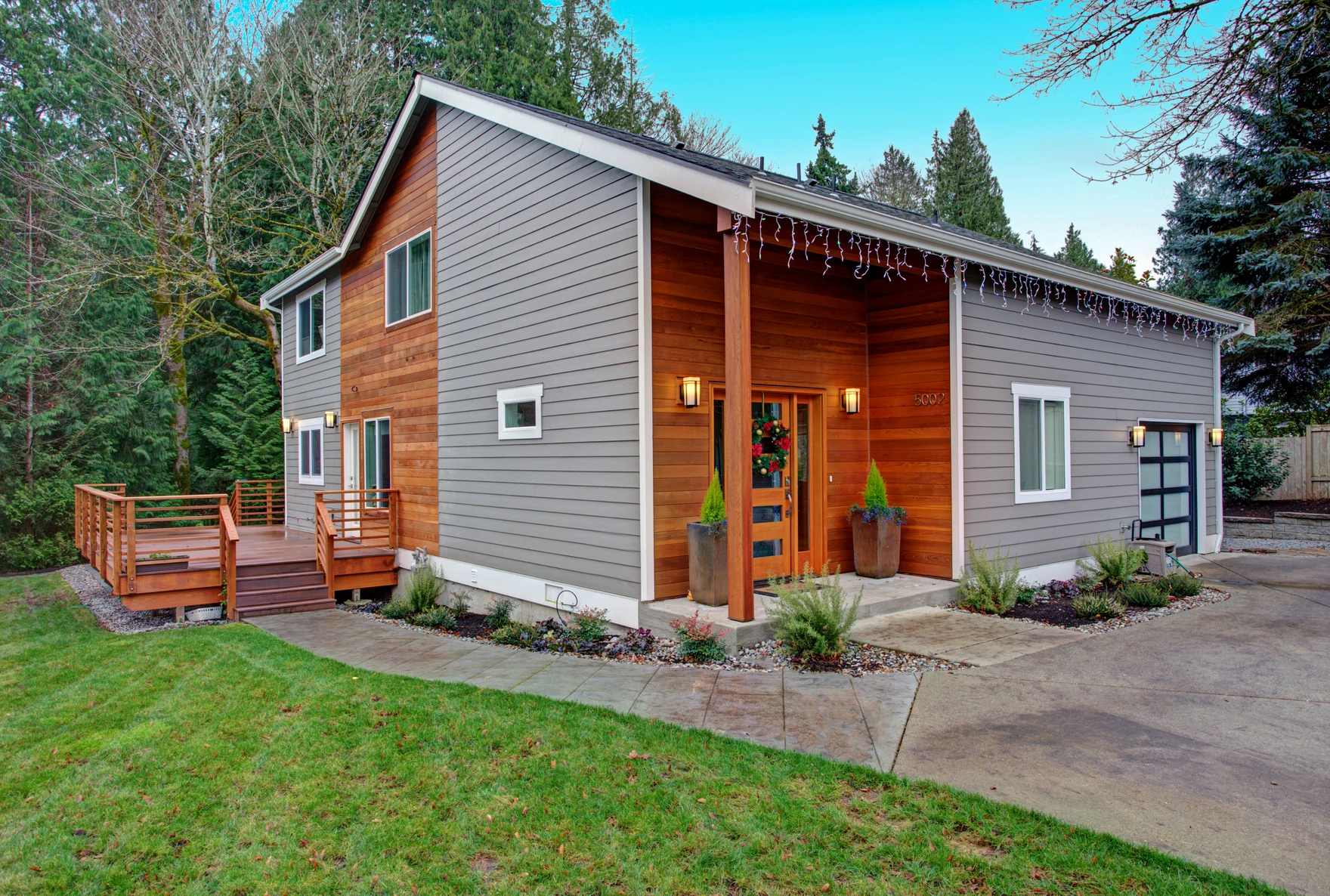 Modern two-story house with gray and wooden siding, festive lights, a deck on the side, and a front door decorated for the holidays, surrounded by trees and a neatly landscaped yard.