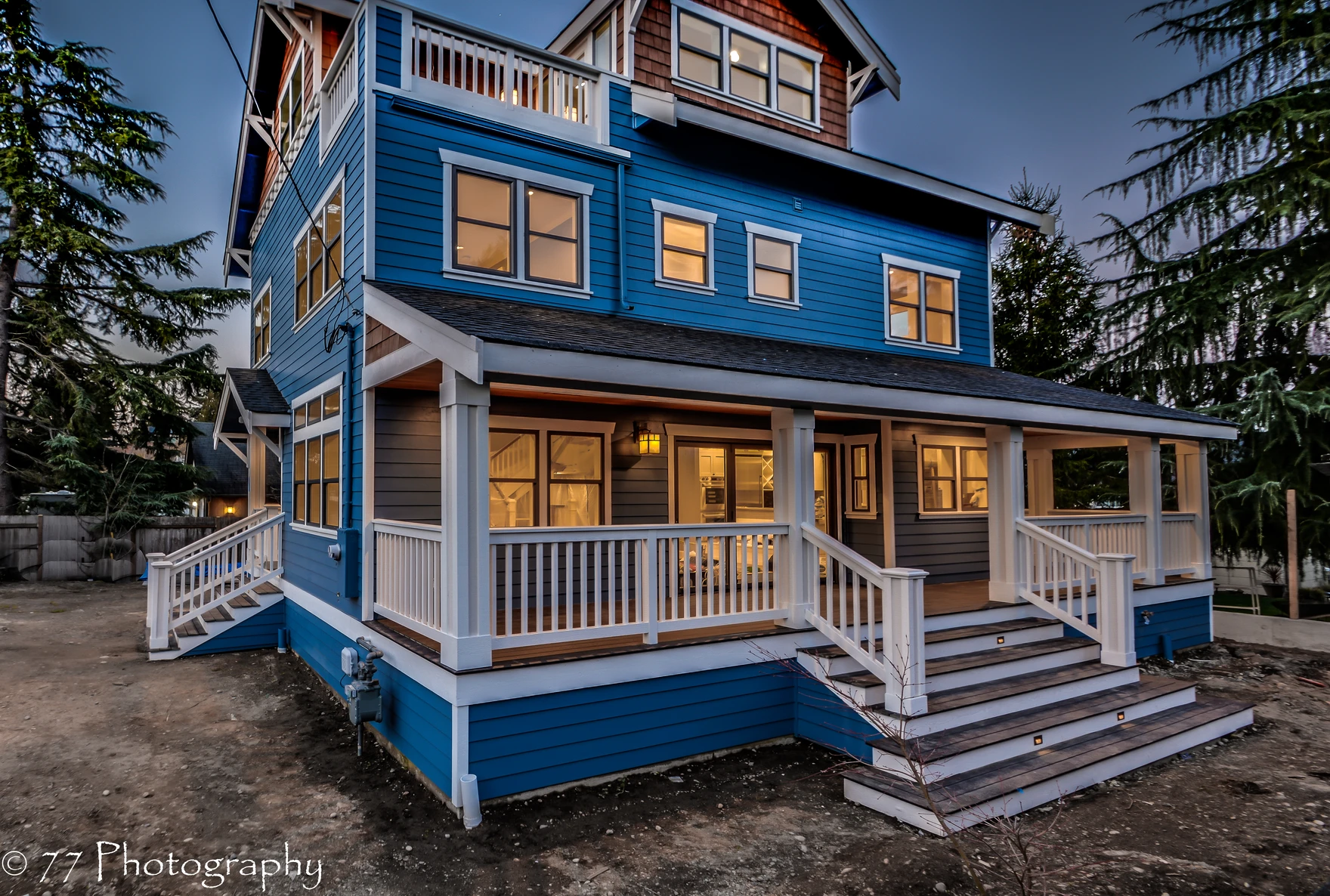 A modern blue three-story house with white trim, lit windows, and a wraparound porch sits nestled in West Seattle. Wide steps lead to the main entrance. Surrounded by trees, it’s dusk outside.