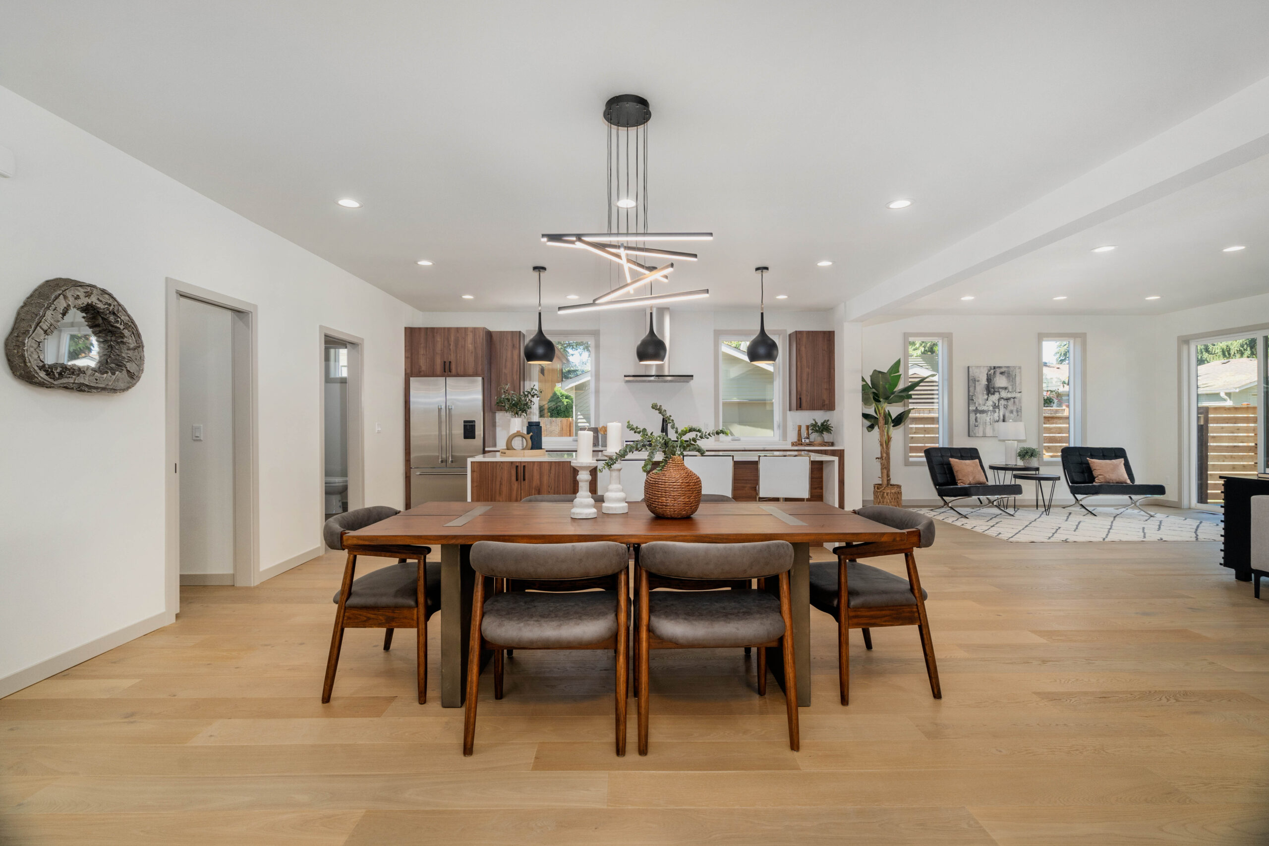 Modern open-concept kitchen and dining area with a wooden table, six chairs, pendant lights, and stylish decor. Living area with large windows and neutral tones visible in the background.