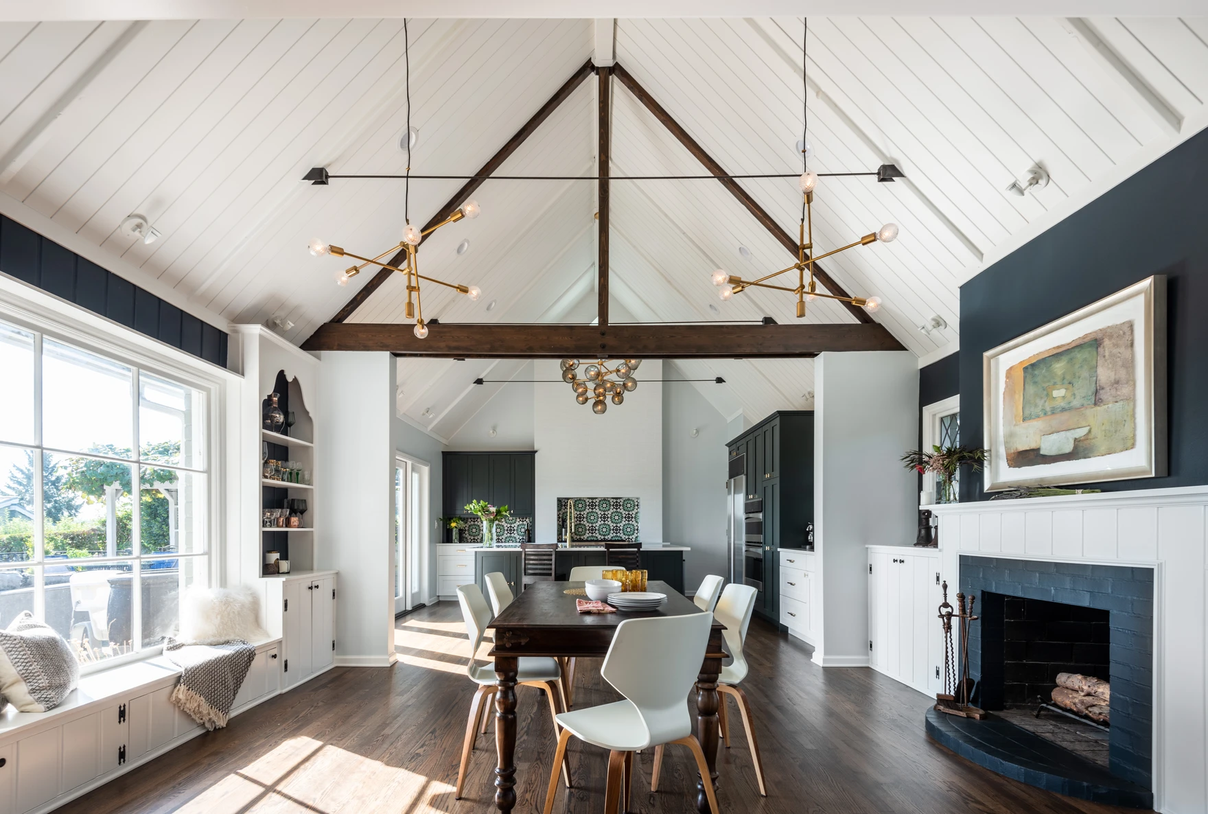 Bright, modern dining room with vaulted ceiling, exposed beams, contemporary light fixtures, large windows, a fireplace with artwork above, and a wooden table with white chairs.